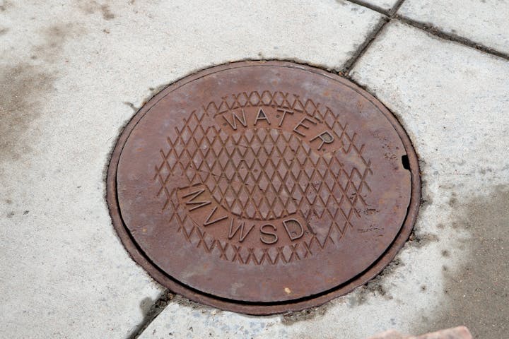 A round, rusty manhole cover labeled "WATER MVWSD" set into concrete pavement.
