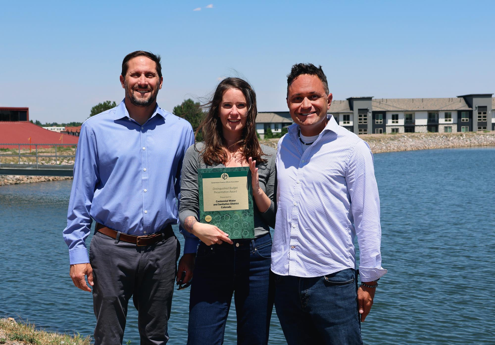Three people standing by a lake, one holding an award certificate, with buildings in the background.