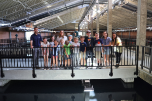 A group of students and three adults stand on a walkway inside a large industrial building with metal beams and overhead lighting.