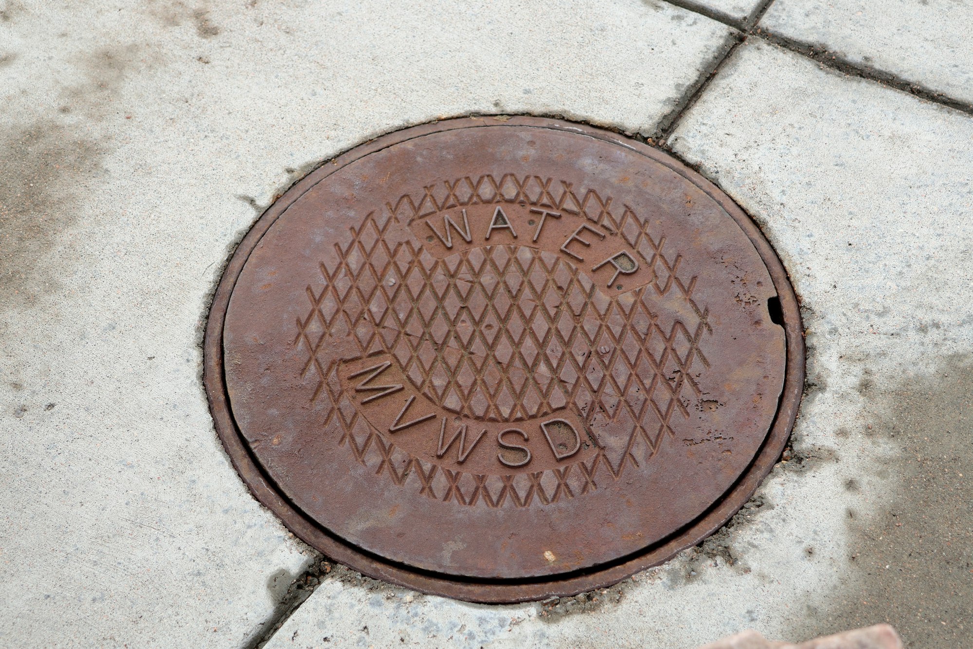 A round, rusty manhole cover labeled "WATER MVWSD" on a concrete sidewalk.