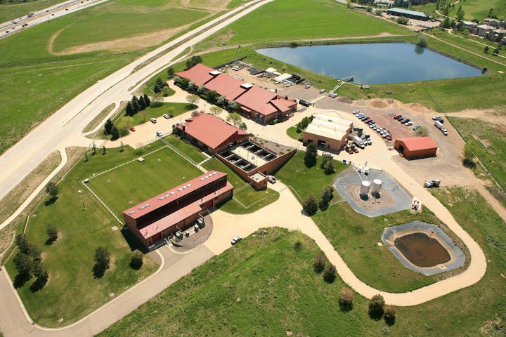 Aerial view of a complex with red-roofed buildings, parking, roads, green areas, a pond, and adjacent highway.