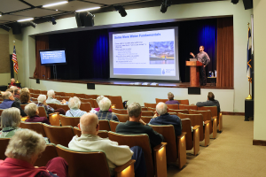 A speaker presents "Basic Water Use Fundamentals" to an audience in an auditorium.