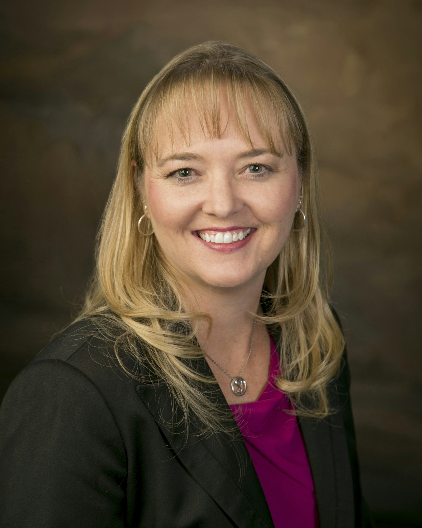 A woman smiling, wearing a black blazer and a pink top, with blonde hair and a neutral background.