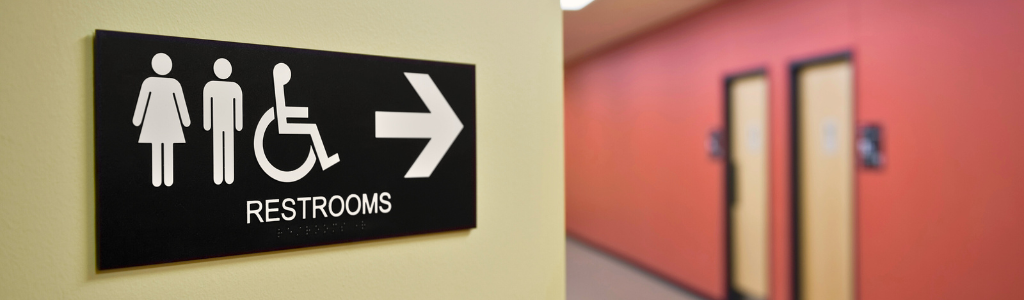 Restroom sign with male, female, and wheelchair symbols, pointing right towards a hallway with red walls.