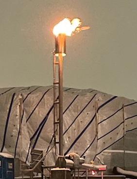 Flaring stack at an industrial site, burning off gas with a bright flame atop a tall chimney, against a twilight sky.