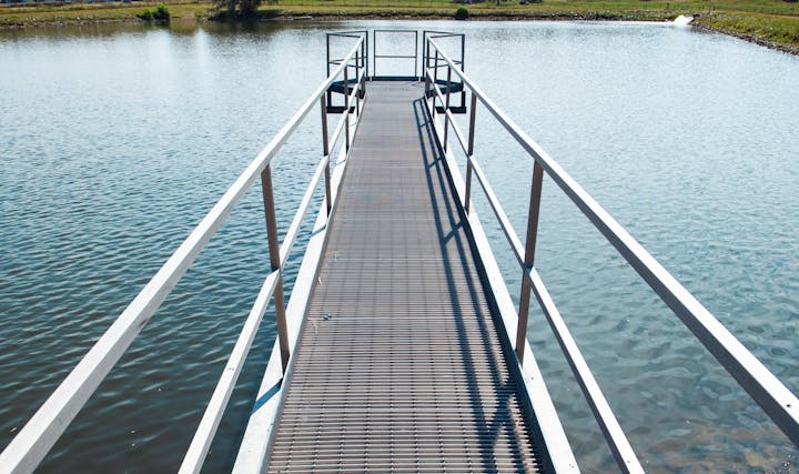 Metal pier extending over a water storage reservoir, with grassy shores in the background.