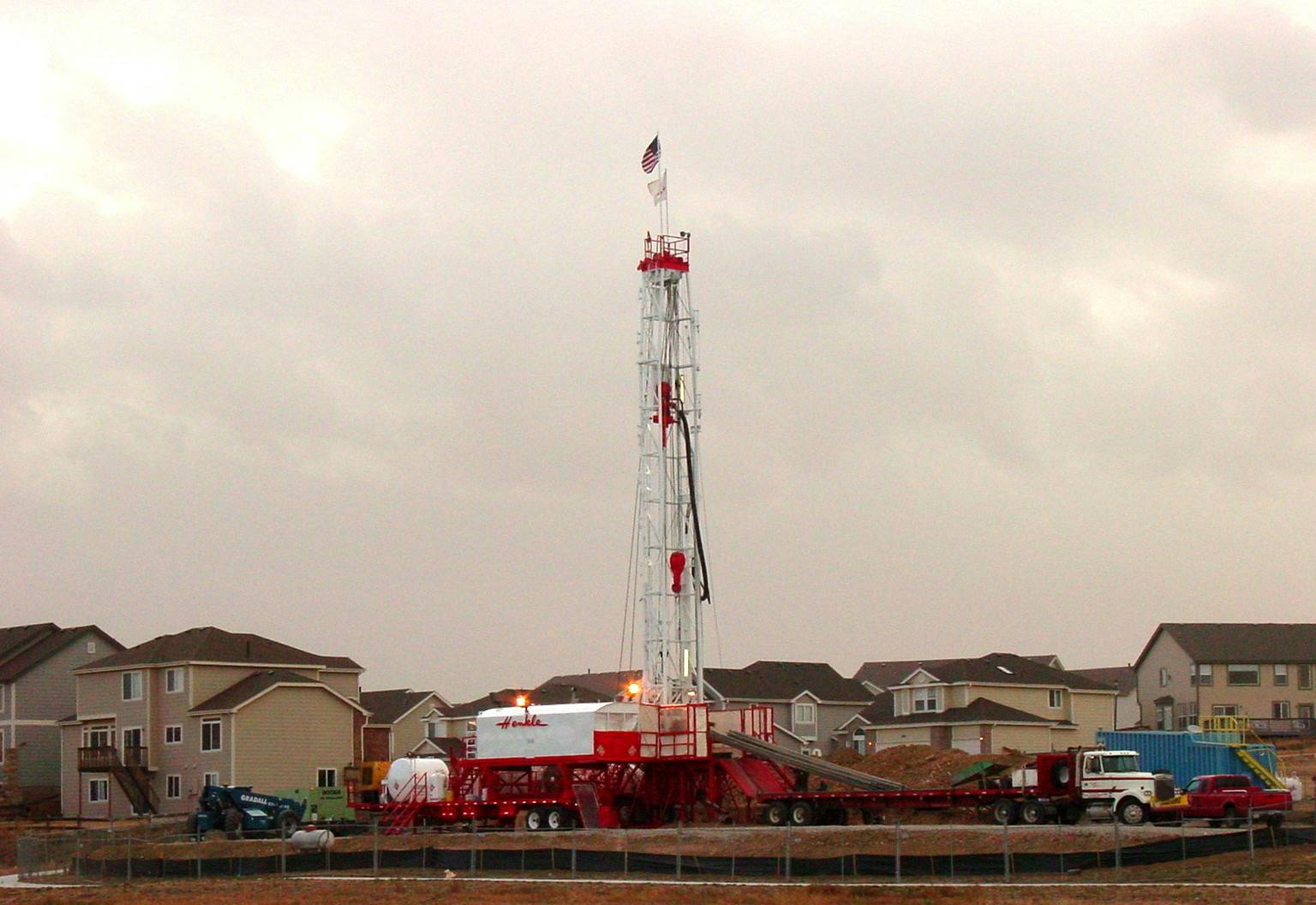A drilling rig is set up among residential houses, with various vehicles and equipment nearby, under a cloudy sky.