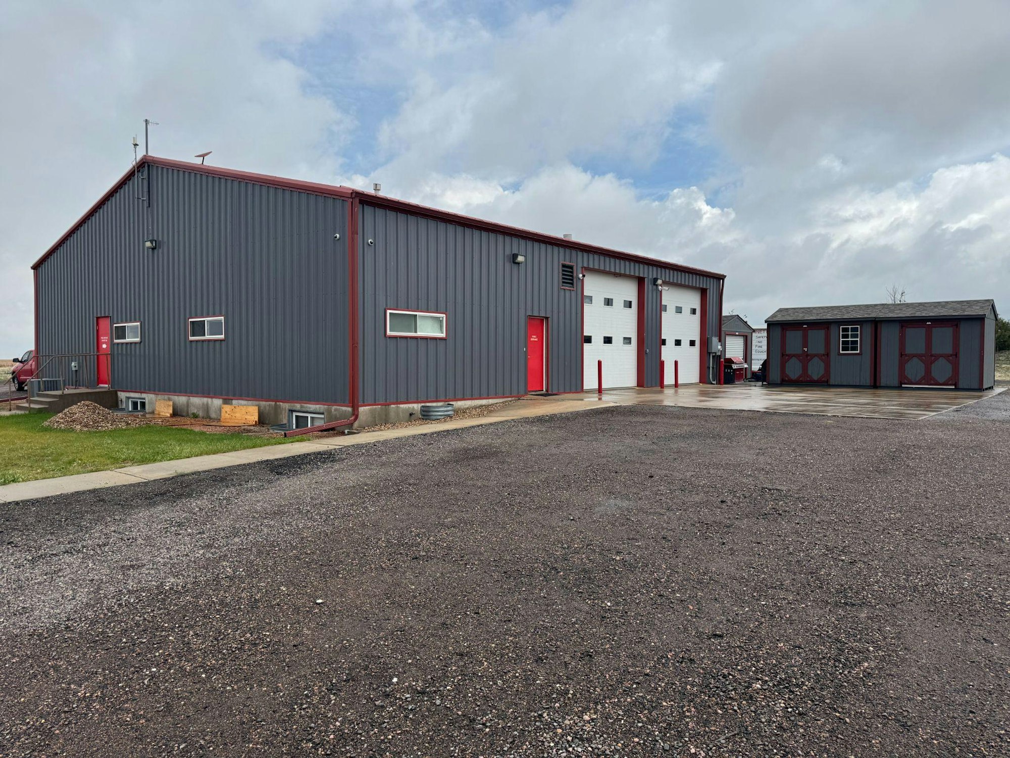 A gray and red industrial building with garage doors, a smaller adjacent shed, on a gravel and concrete lot under a cloudy sky.