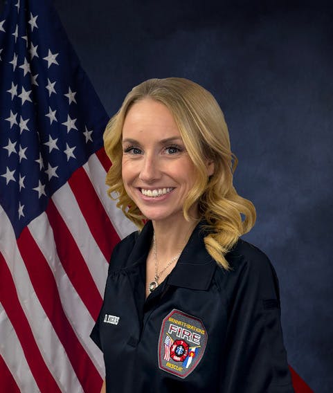 A smiling woman in a fire department uniform stands in front of an American flag.