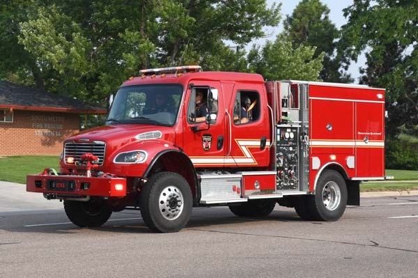 A red fire truck on the road with visible equipment and green trees in the background.
