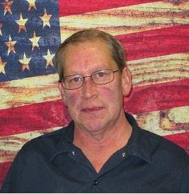 The image features a man in glasses standing in front of an American flag backdrop.