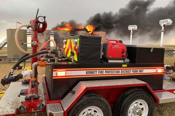 A foam unit from Bennett Fire Protection District near burning tanks, emitting flames and thick smoke into the sky.