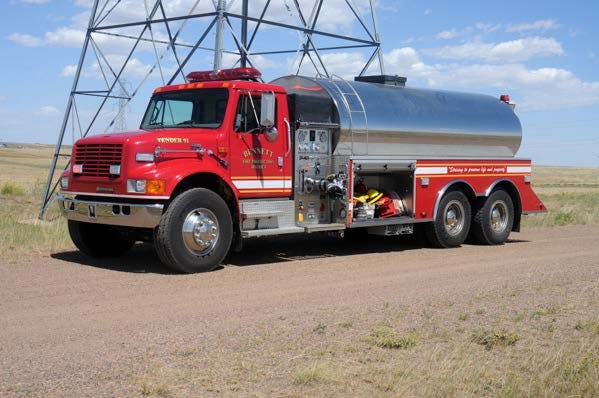 A red fire tanker truck parked on a dirt road beside a field, with a metal structure in the background.