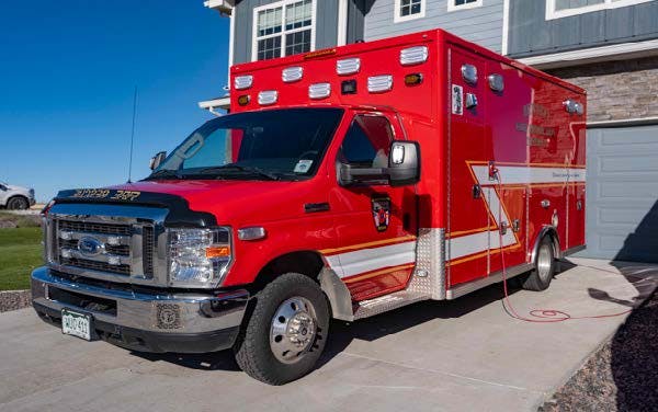 A red ambulance is parked in a driveway next to a house, with a blue sky in the background.