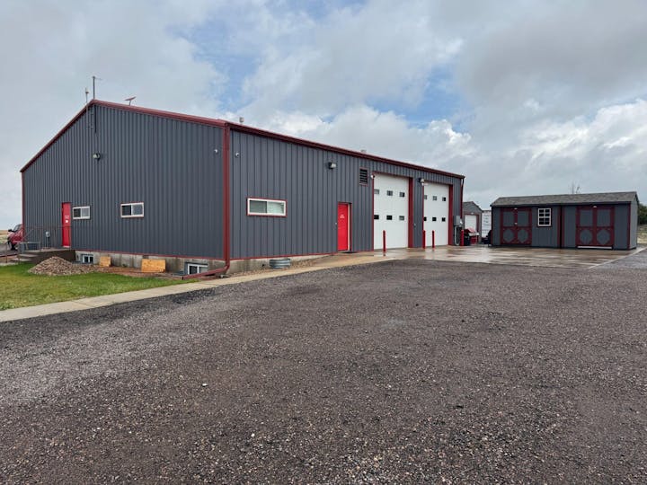 Two gray buildings with red accents, a larger industrial style with garage doors and a smaller one, on a gravel and concrete lot.