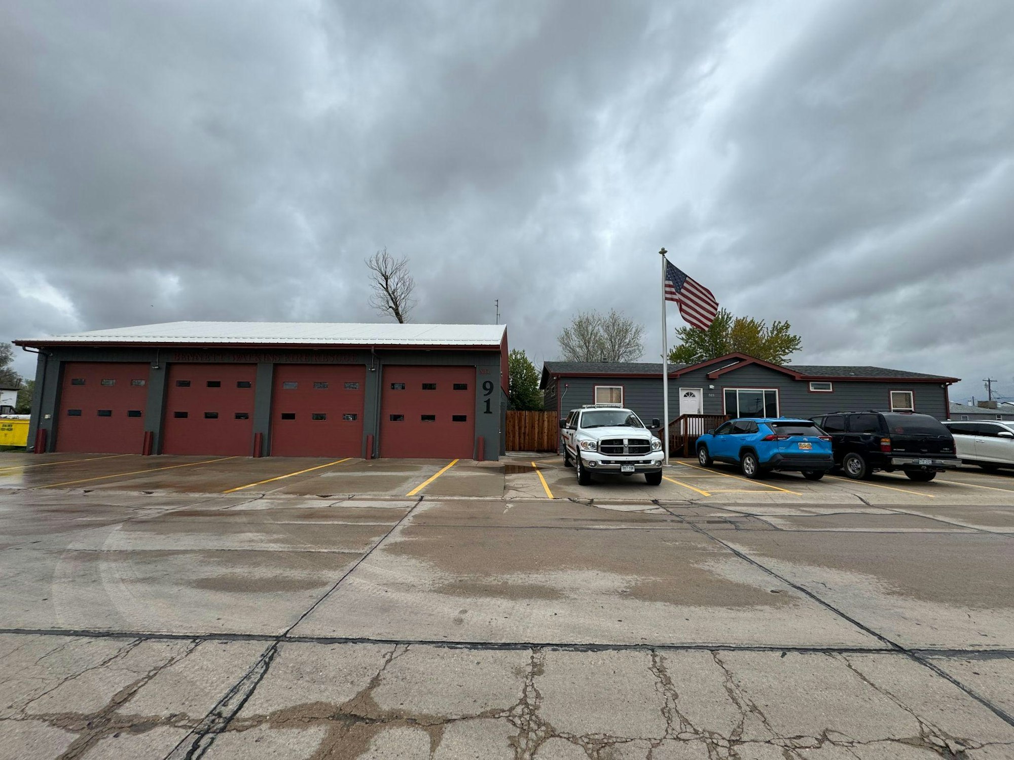 A fire station with red garage doors, several parked cars, and an American flag outside, under a cloudy sky.