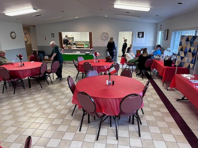 A room with red-covered tables, people sitting and standing, and a kitchen area in the background.