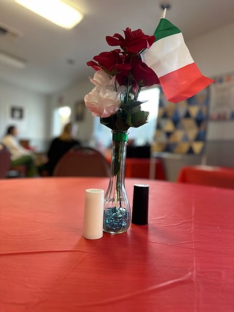 A vase with red and white flowers and an Italian flag on a red tablecloth with candles, indoors.