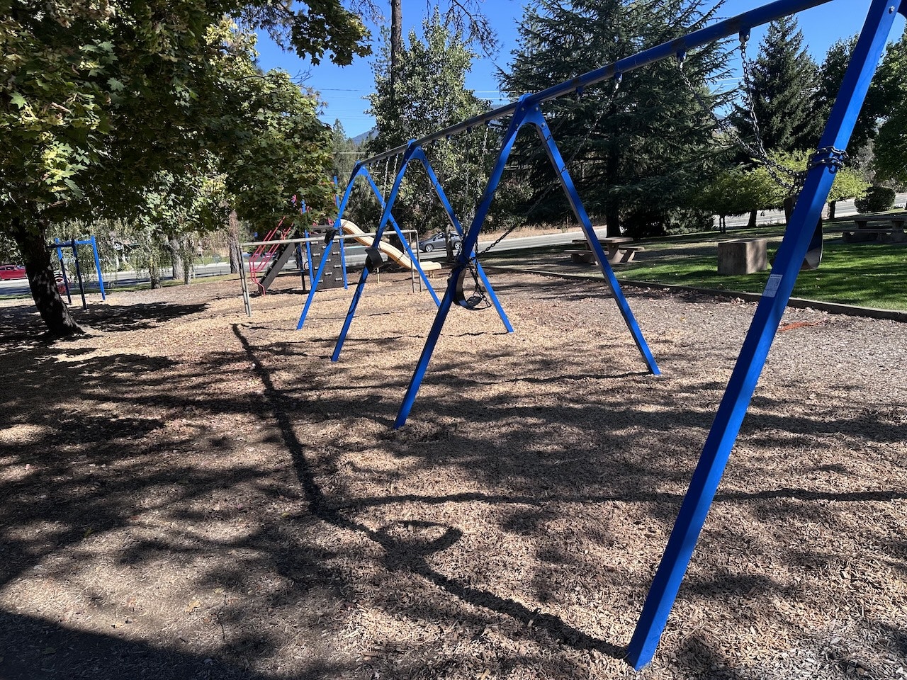 A playground with blue swings, a slide, and shadows under trees on a sunny day.
