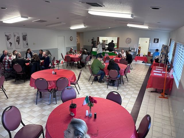 People seated at round tables with red tablecloths in a room, likely at a social gathering or event.