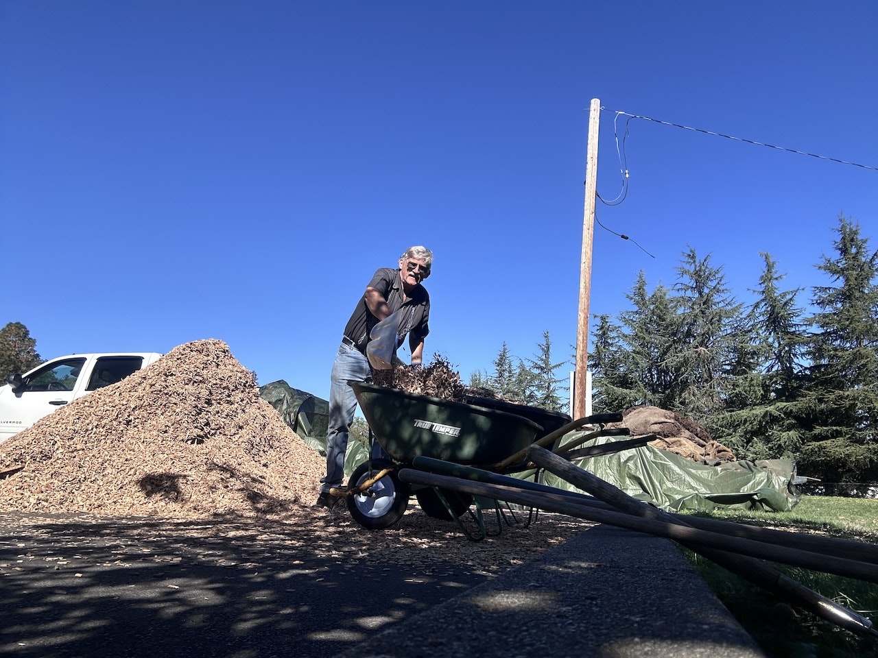 A person shoveling mulch into a wheelbarrow on a sunny day with trees and a pile of mulch in the background.