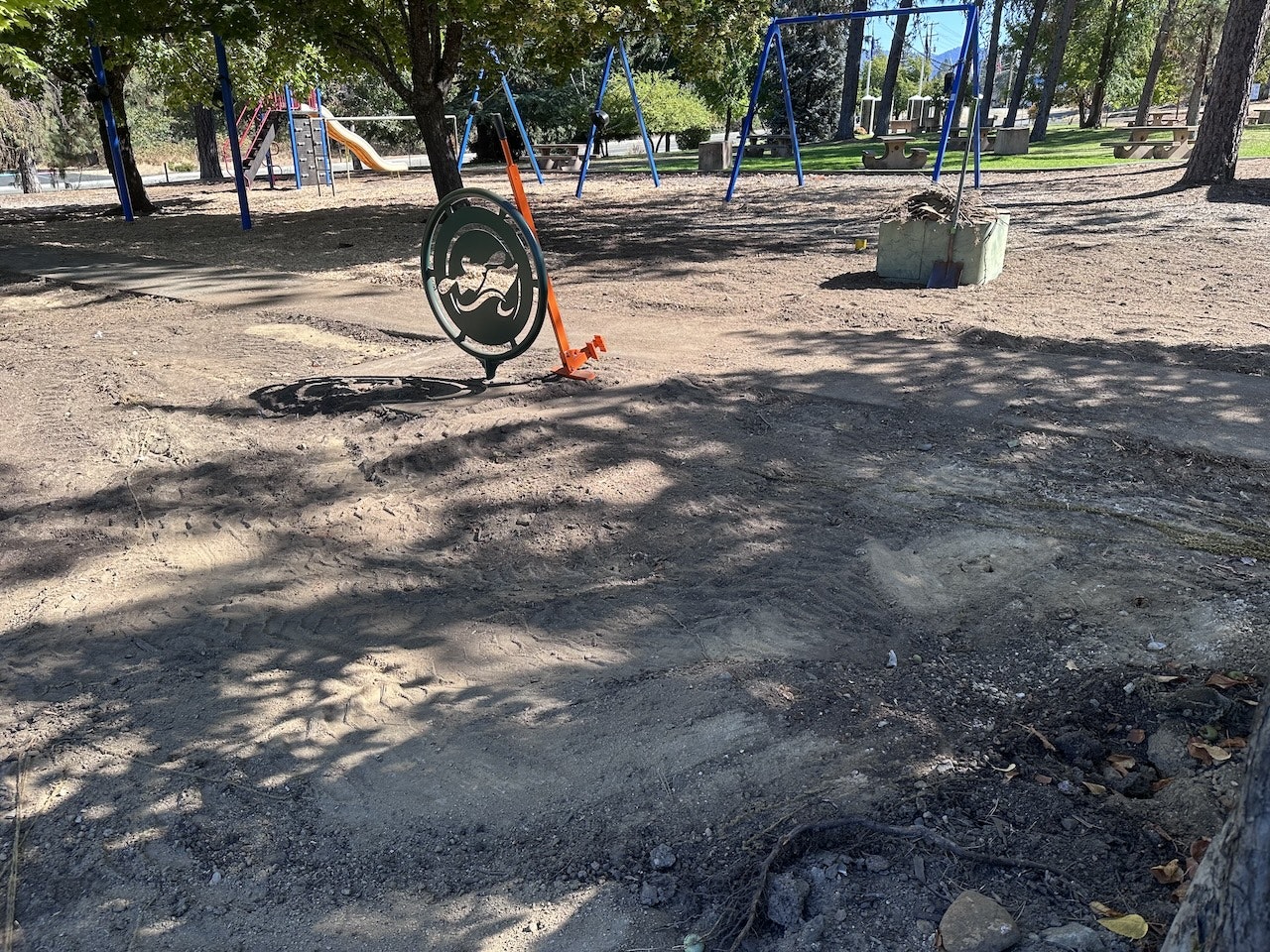 A playground with swings, slide, and a round swing, cast in shadow on sandy ground.