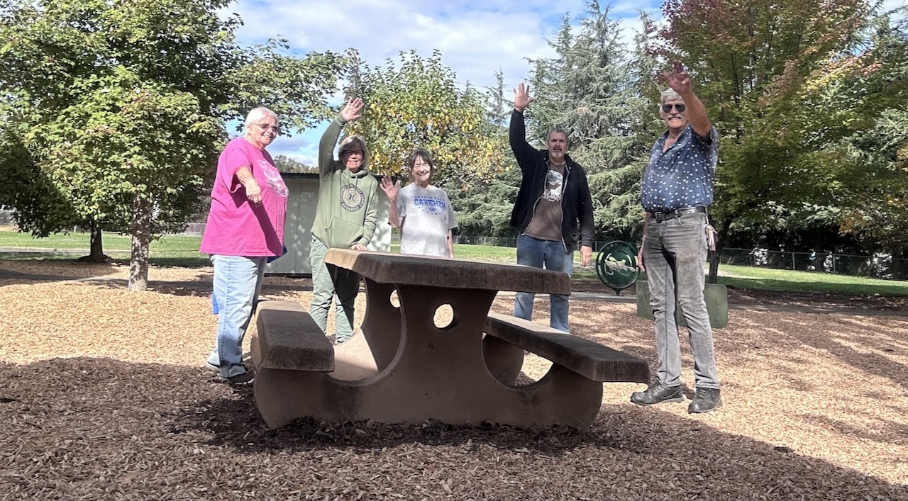 Group of five people waving and standing behind a concrete table in a park, trees and clear sky in the background.