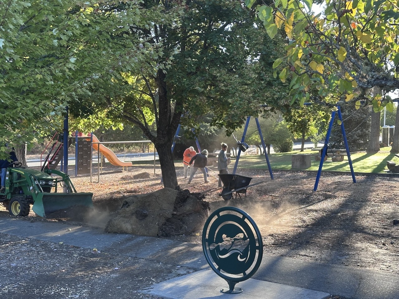 Playground with workers, a tractor, swings, slide, and dust in the air. A decorative sign in the foreground.