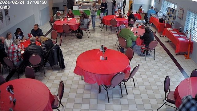 An indoor gathering with people sitting at tables covered with red cloths.