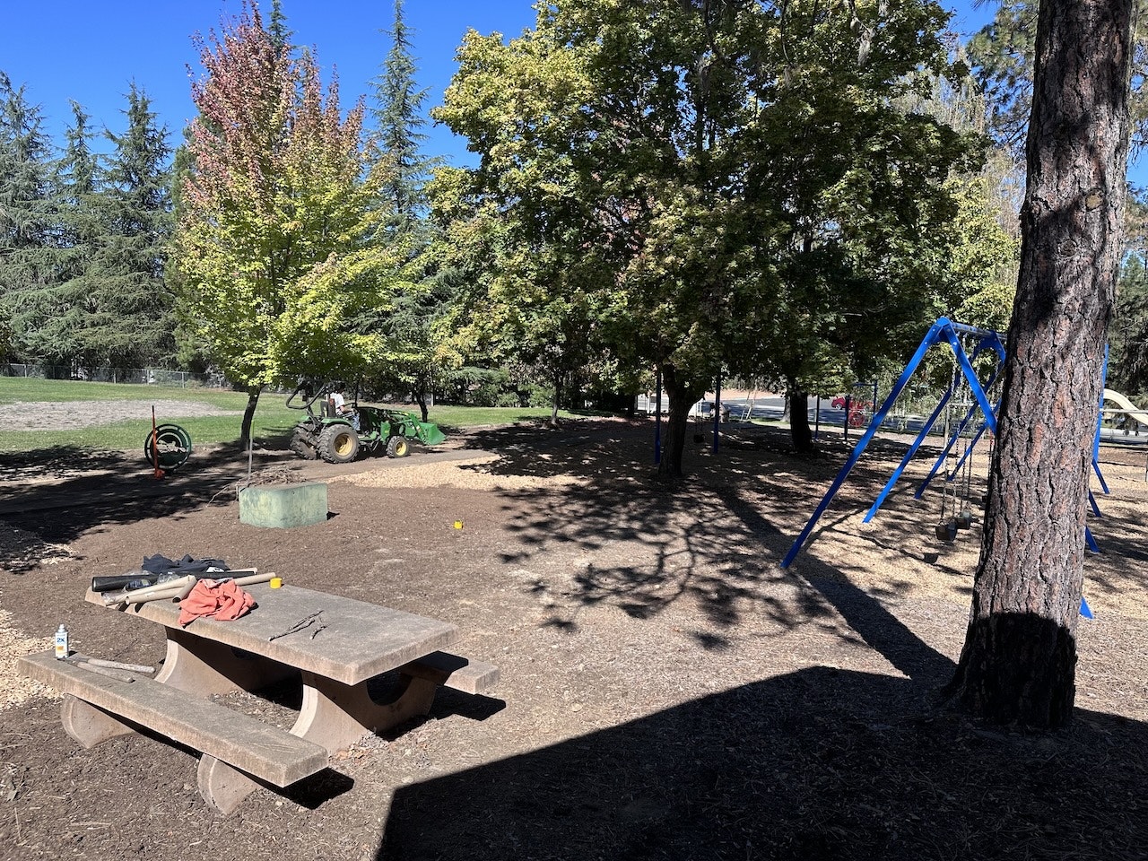 A playground with swings, a table, trees, and a tractor in the background.