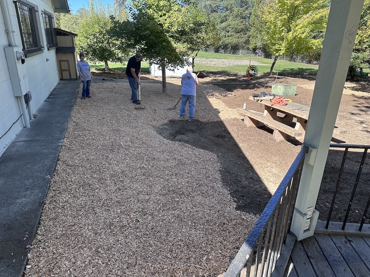 Three people raking and spreading mulch in an outdoor area with trees, a picnic table, and a wheelbarrow.