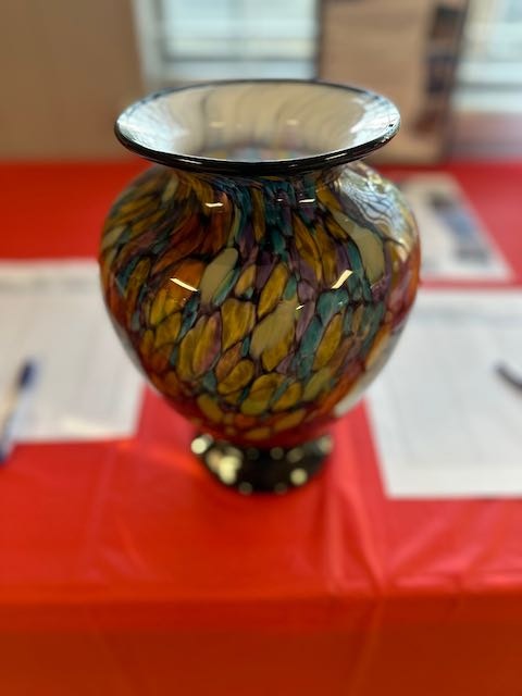 A colorful glass vase on a red table, with blurred papers and pens in the background.
