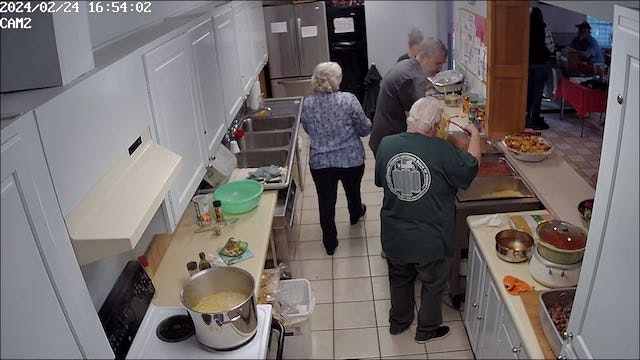 A group of people cooking and serving food in a kitchen with a security camera timestamp.