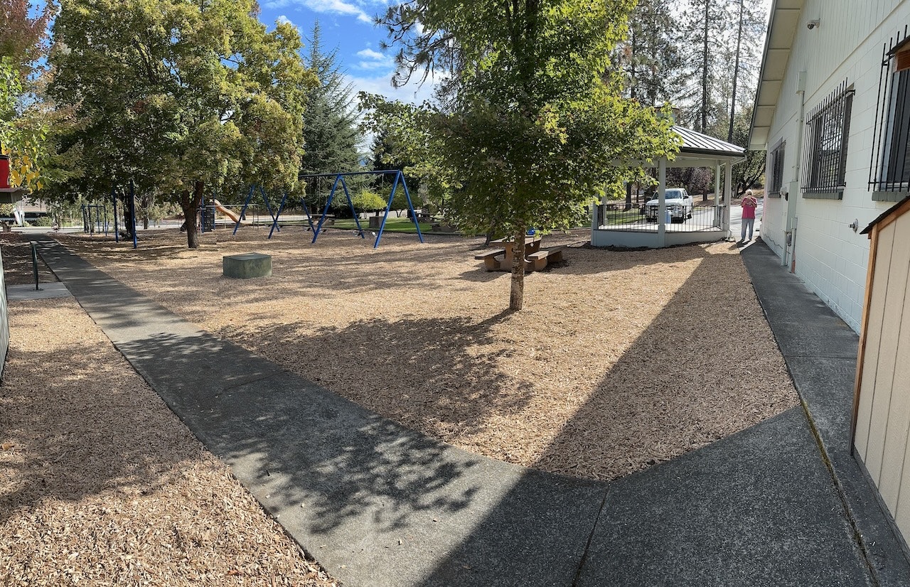 A playground with swings, benches, trees, and a person in the distance.