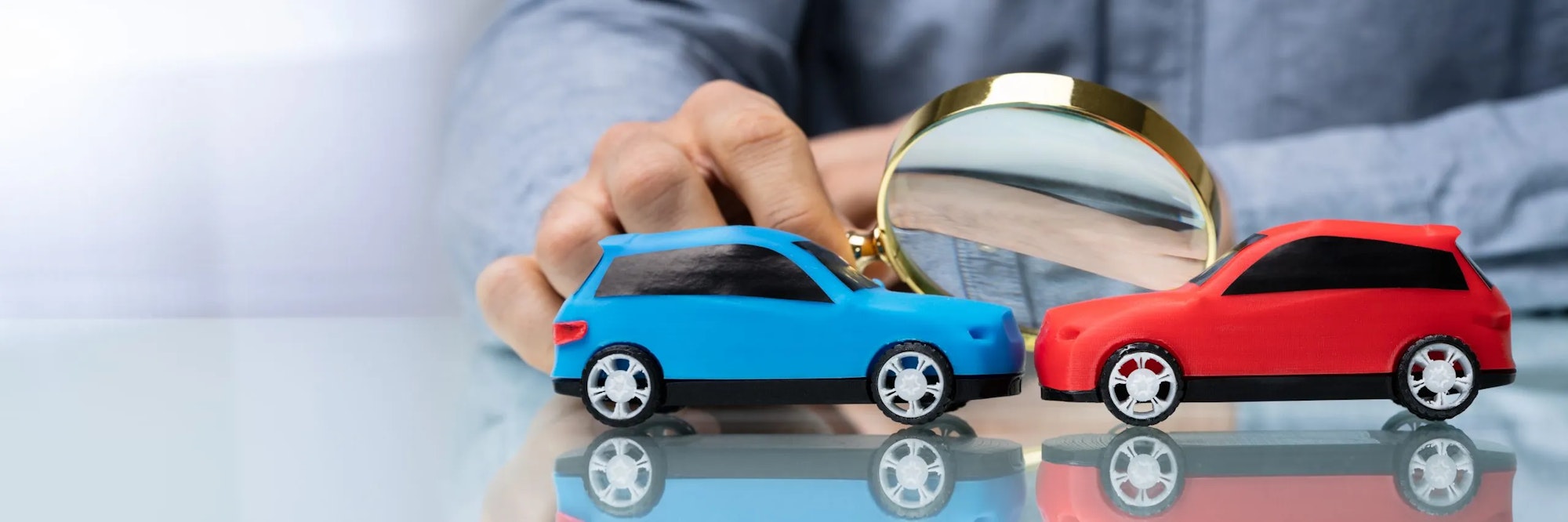 A hand holding a magnifying glass over two toy cars, one blue and one red.
