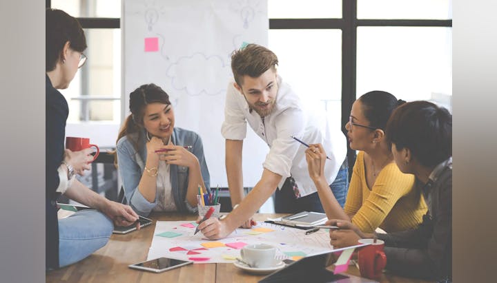 A diverse group of people collaboratively working around a table with papers and markers, in a bright office setting.