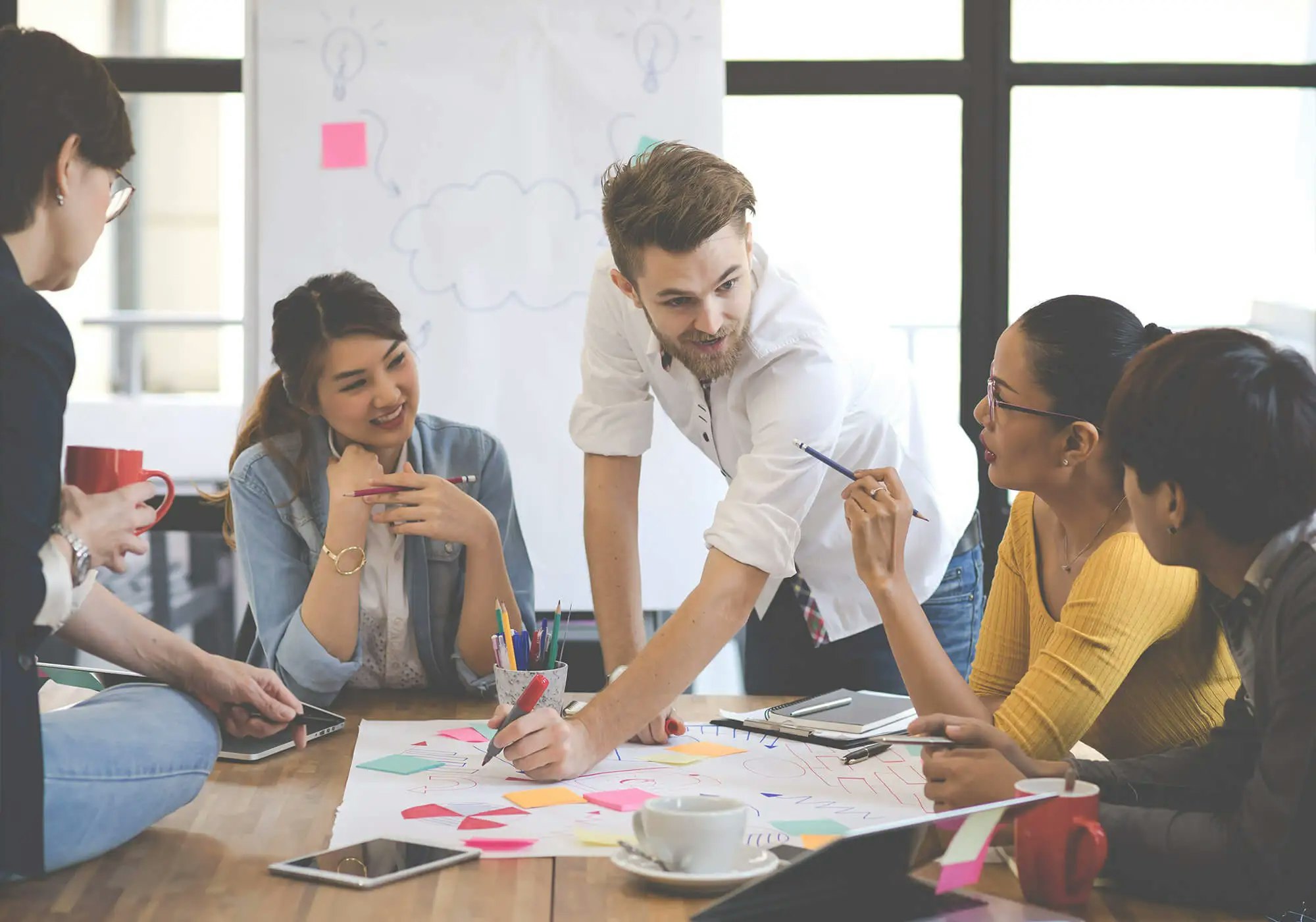 A diverse group of people collaboratively working around a table with papers and markers, in a bright office setting.