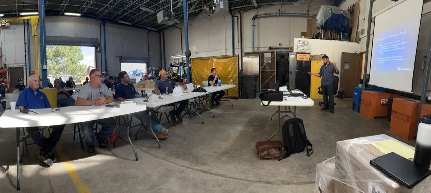 A group attending a presentation in a workshop setting with a speaker, tables, chairs, and a projection screen.