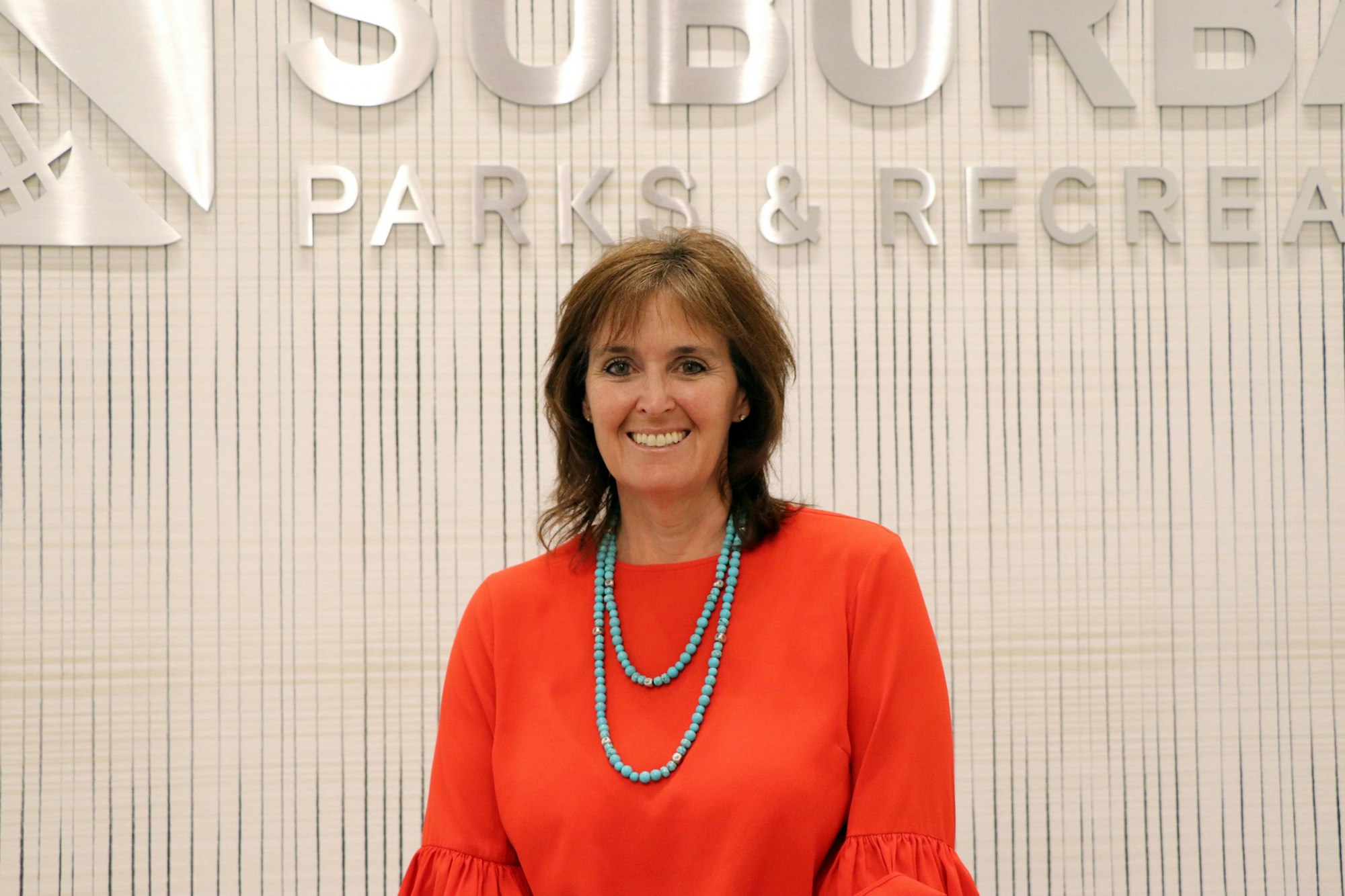 Woman in orange blouse with turquoise necklace, smiling in front of "SUBURBAN PARKS & RECREATION" sign.
