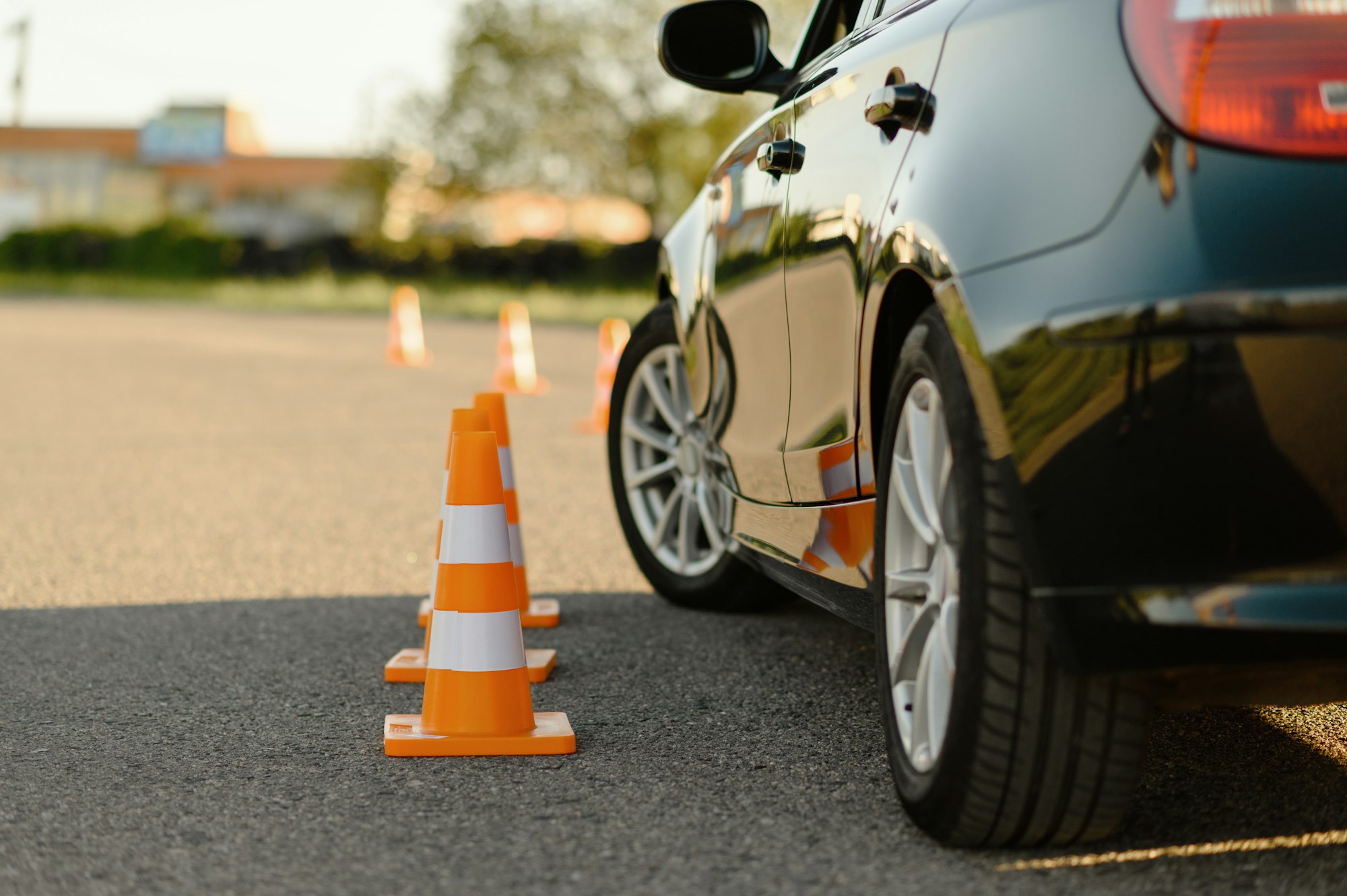 A black car is parked near a line of orange and white traffic cones on an asphalt surface, likely for a driving practice session.