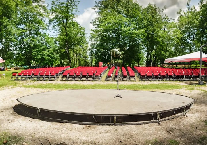 Outdoor stage with microphone and red chairs arranged for an audience, surrounded by trees.