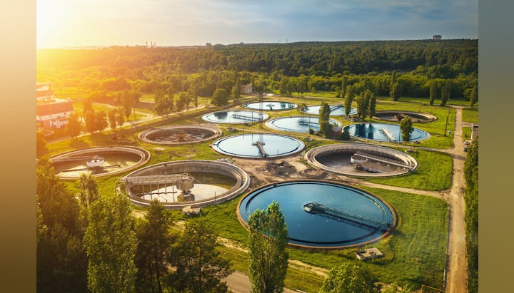 Aerial view of a water treatment plant with circular tanks, surrounded by greenery and trees.