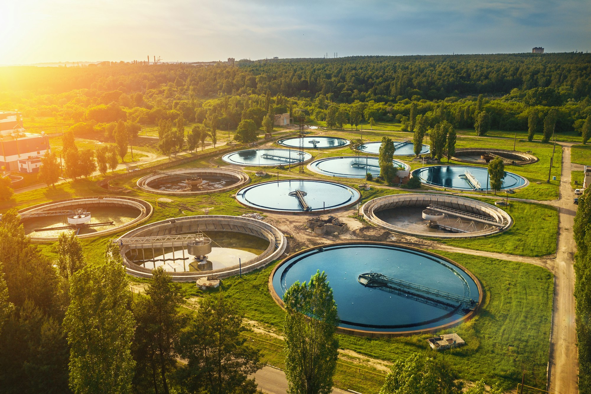 Aerial view of a water treatment plant with circular tanks, surrounded by greenery and trees.