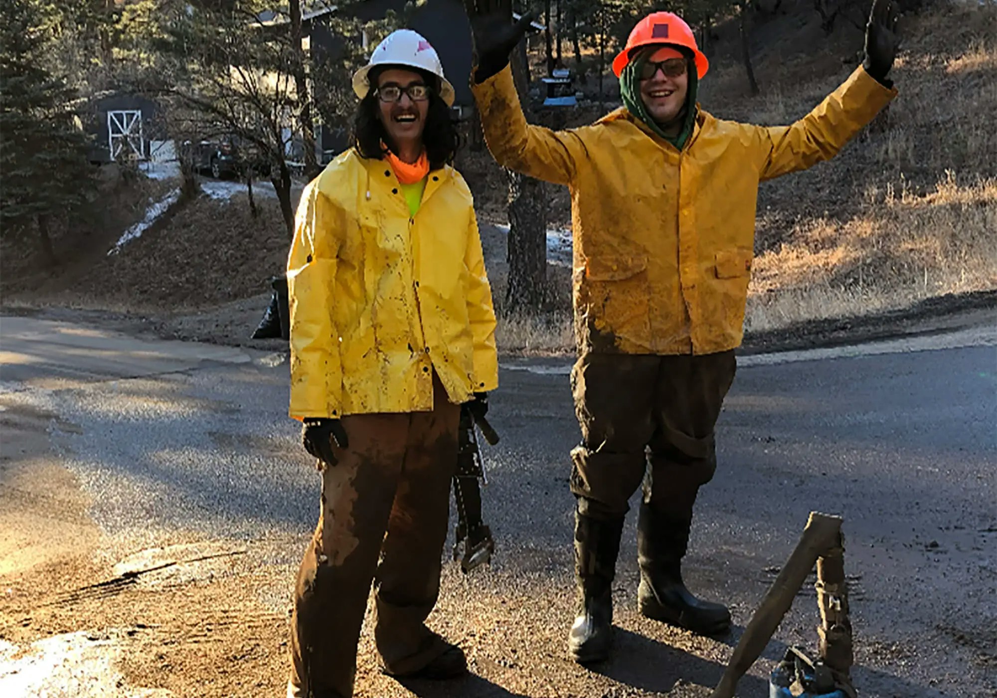 Two people in hi-vis gear and hard hats smiling and waving, with a forested background.