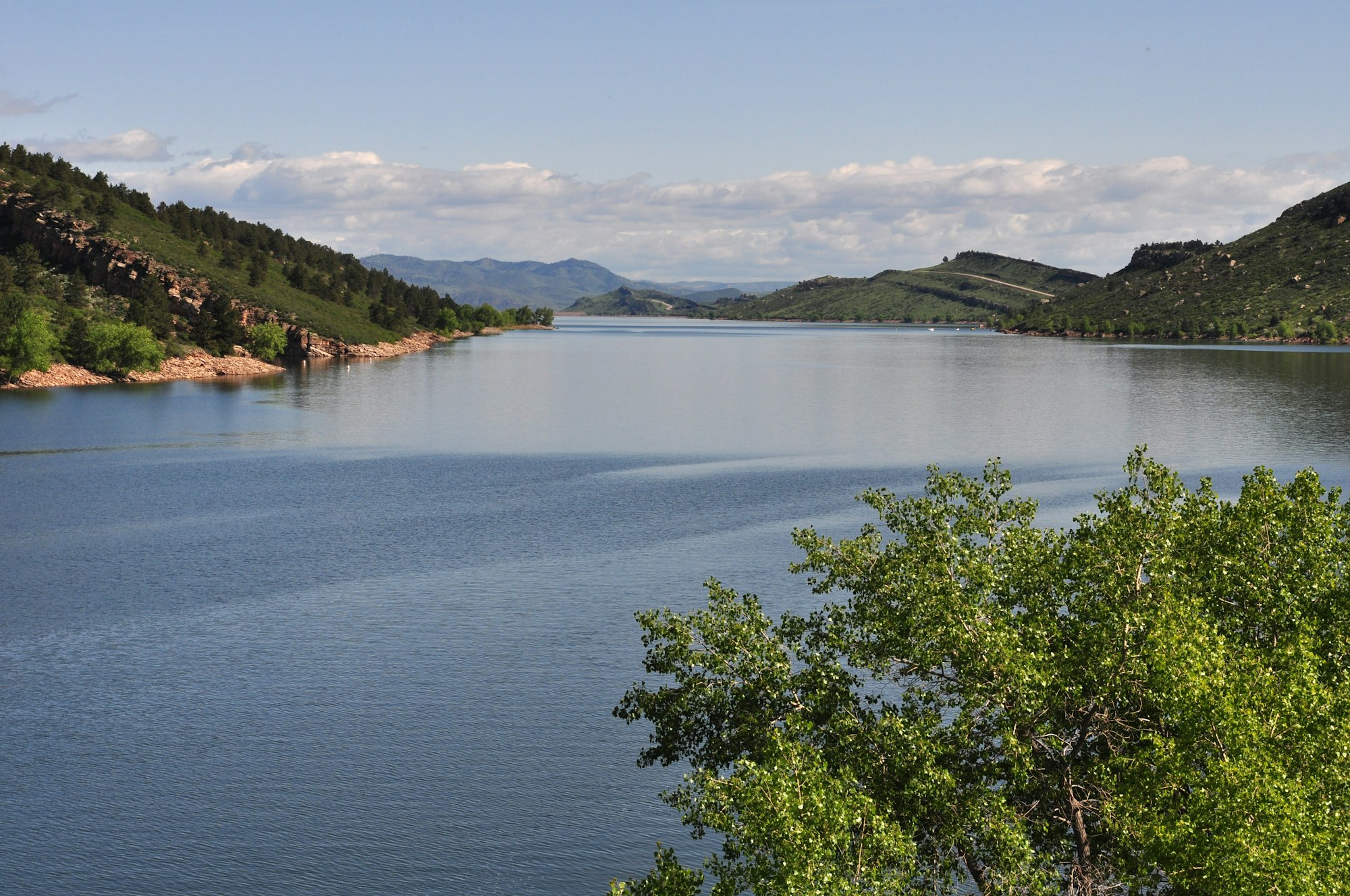 A calm lake surrounded by green hills and trees under a partly cloudy sky, reflecting the serene landscape.
