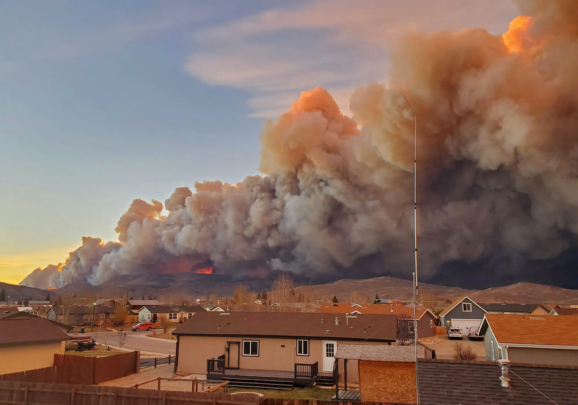 A massive wildfire looms over a residential area with thick smoke and visible flames.