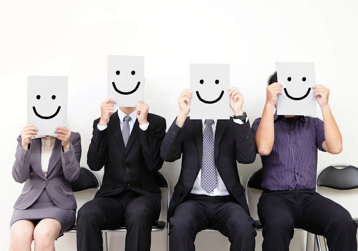 Four people in business attire holding smiley face papers in front of their faces.
