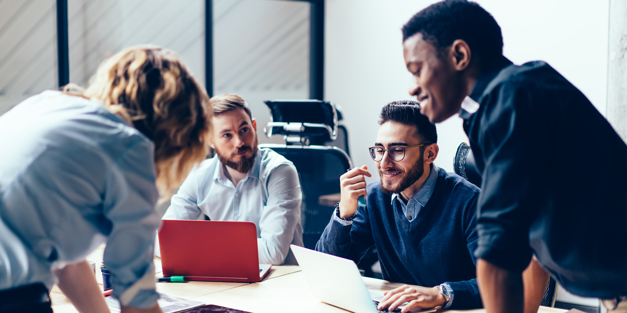 A group of four people having a discussion around a table with laptops in a modern office setting.