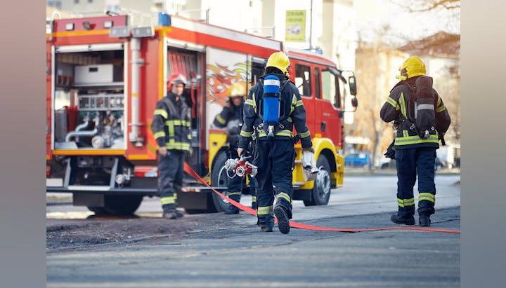 Firefighters in gear approaching an emergency with a fire truck in the background.