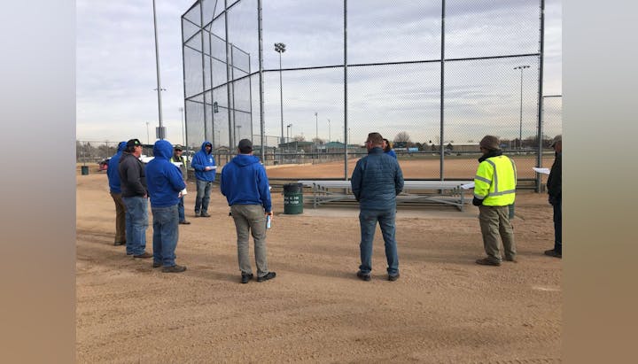 Group of individuals standing near a baseball diamond fence and bleachers.